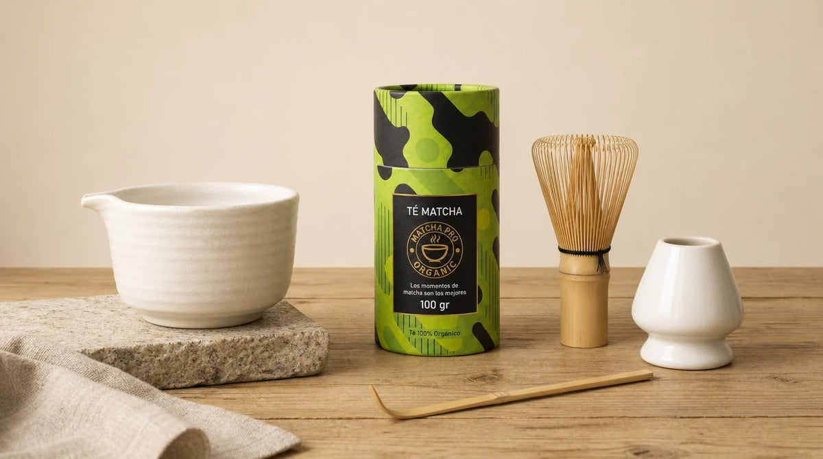 Bote de té matcha MatchaPro de 100 g junto a un chawan de cerámica blanca, un chasen de bambú, un sostenedor blanco y un chashaku sobre una mesa de madera
